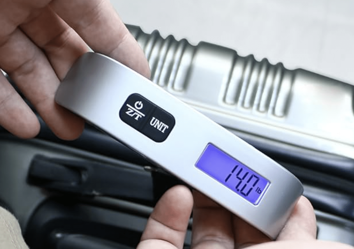 A person holds a digital luggage scale displaying a weight of 14.0 lbs above a silver suitcase.