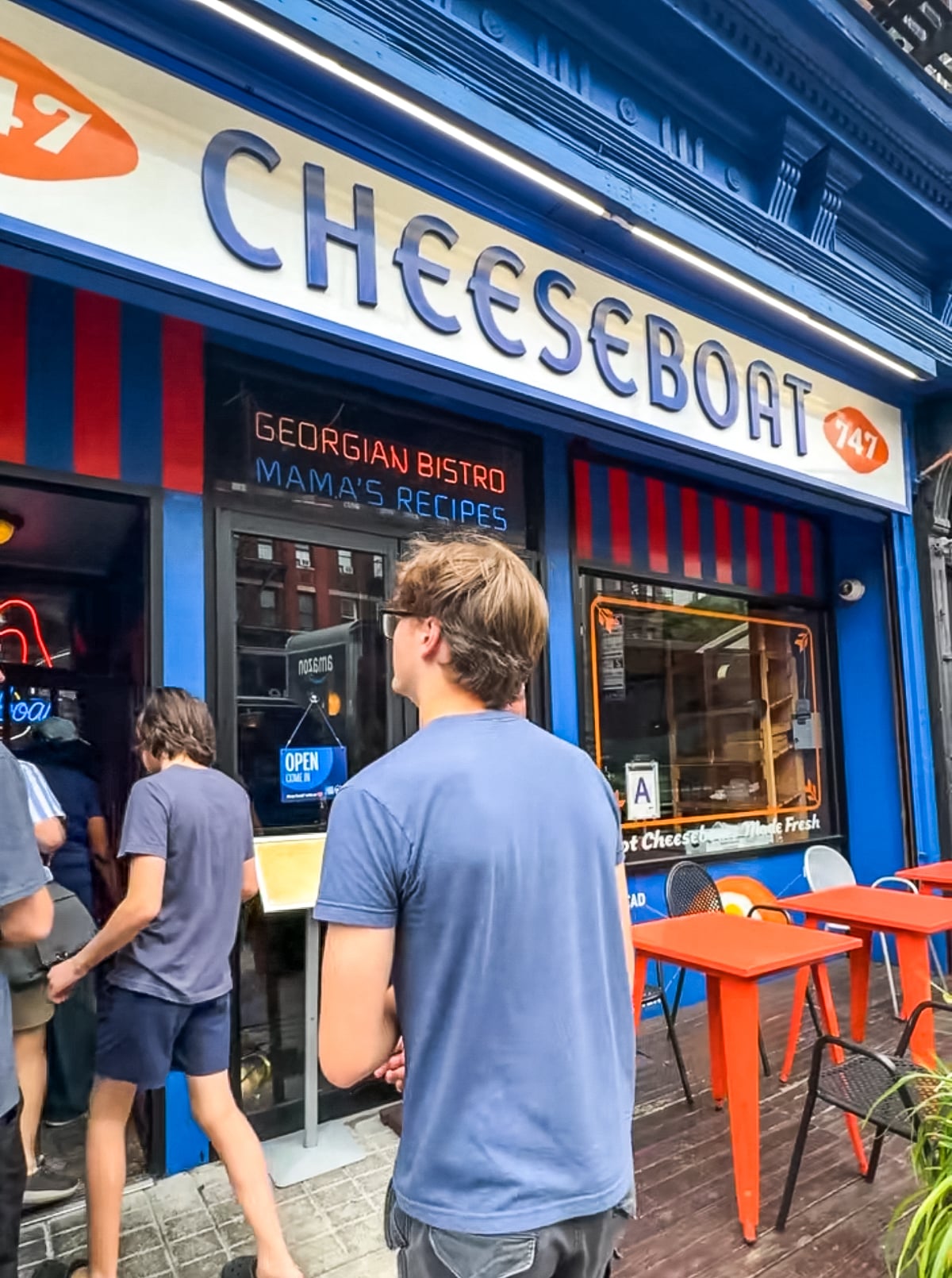 People stand outside the entrance of a blue restaurant called "CHEESEBOAT," which serves Georgian food, with outdoor seating and a sign reading "MAMA'S RECIPES.