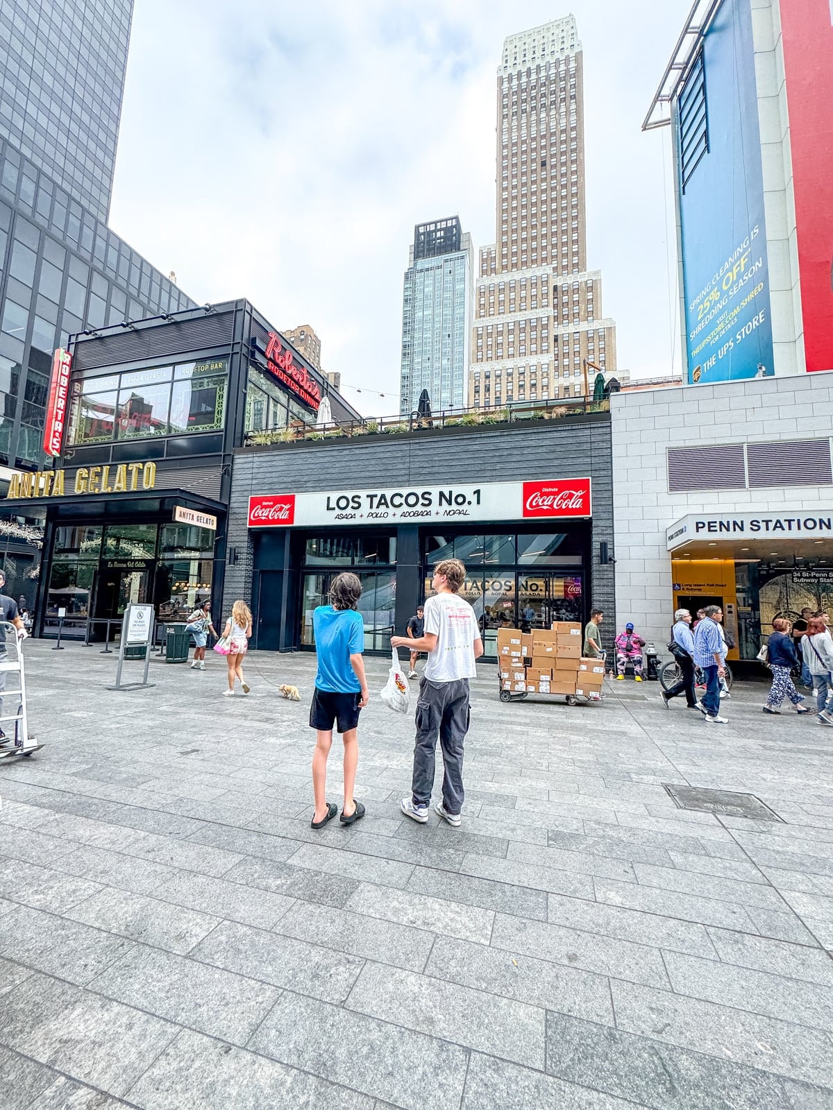 People walk in a plaza outside the Los Tacos No. 1 restaurant, with tall buildings and Penn Station visible in the background.