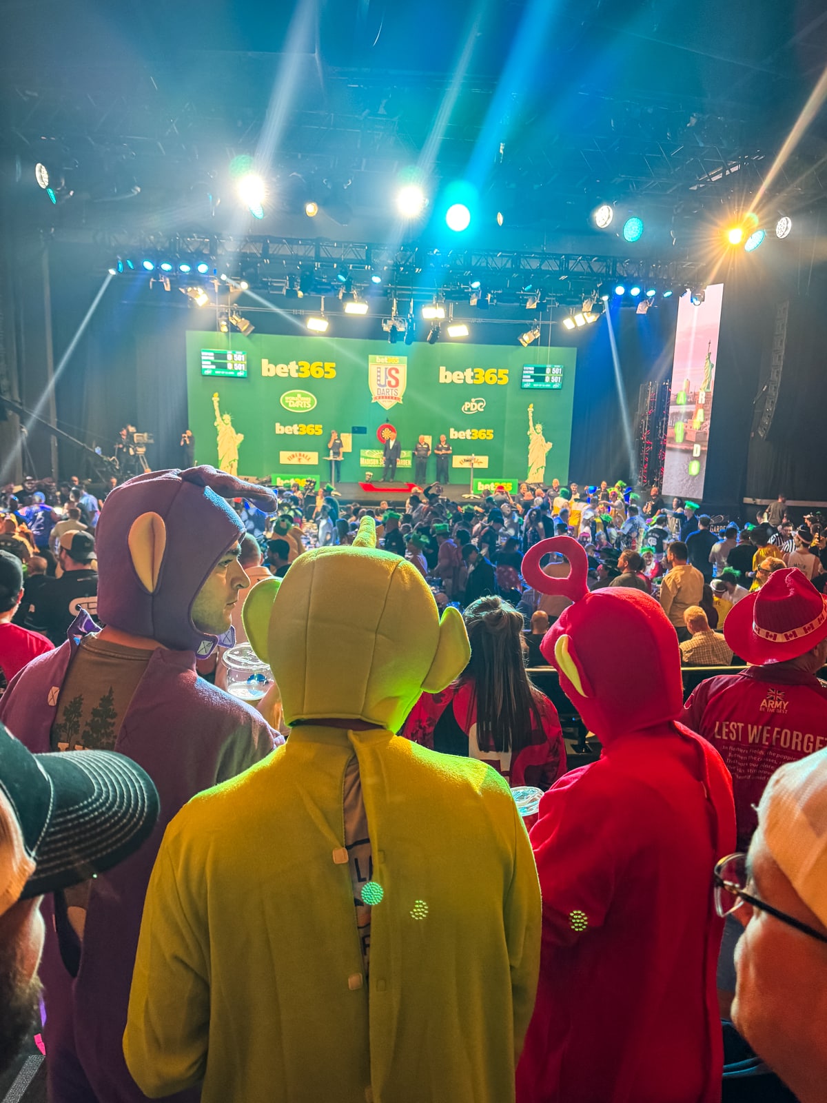 A crowded event with people in colorful costumes, including Teletubbies, facing a stage with bet365 branding and green screens under bright lights.