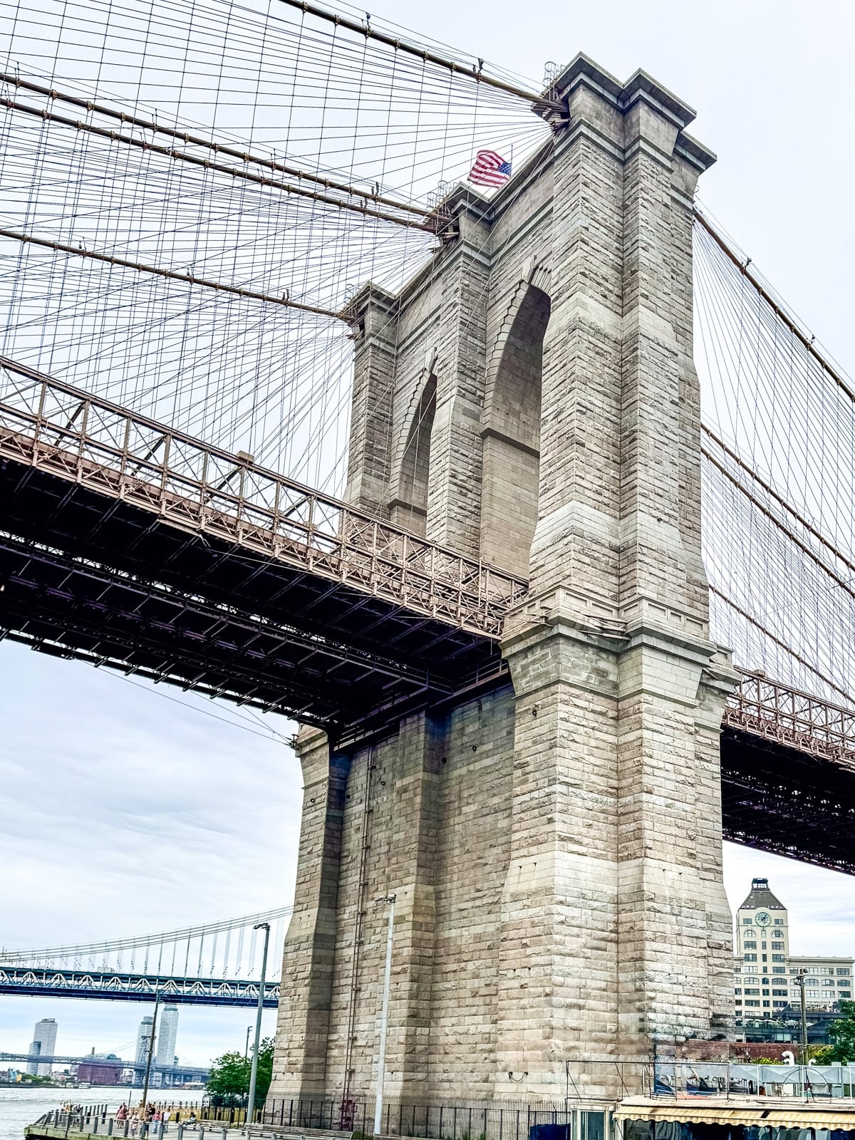 View of the Brooklyn Bridge&rsquo;s stone tower and cables, with the Manhattan Bridge and buildings visible in the background on a cloudy day.