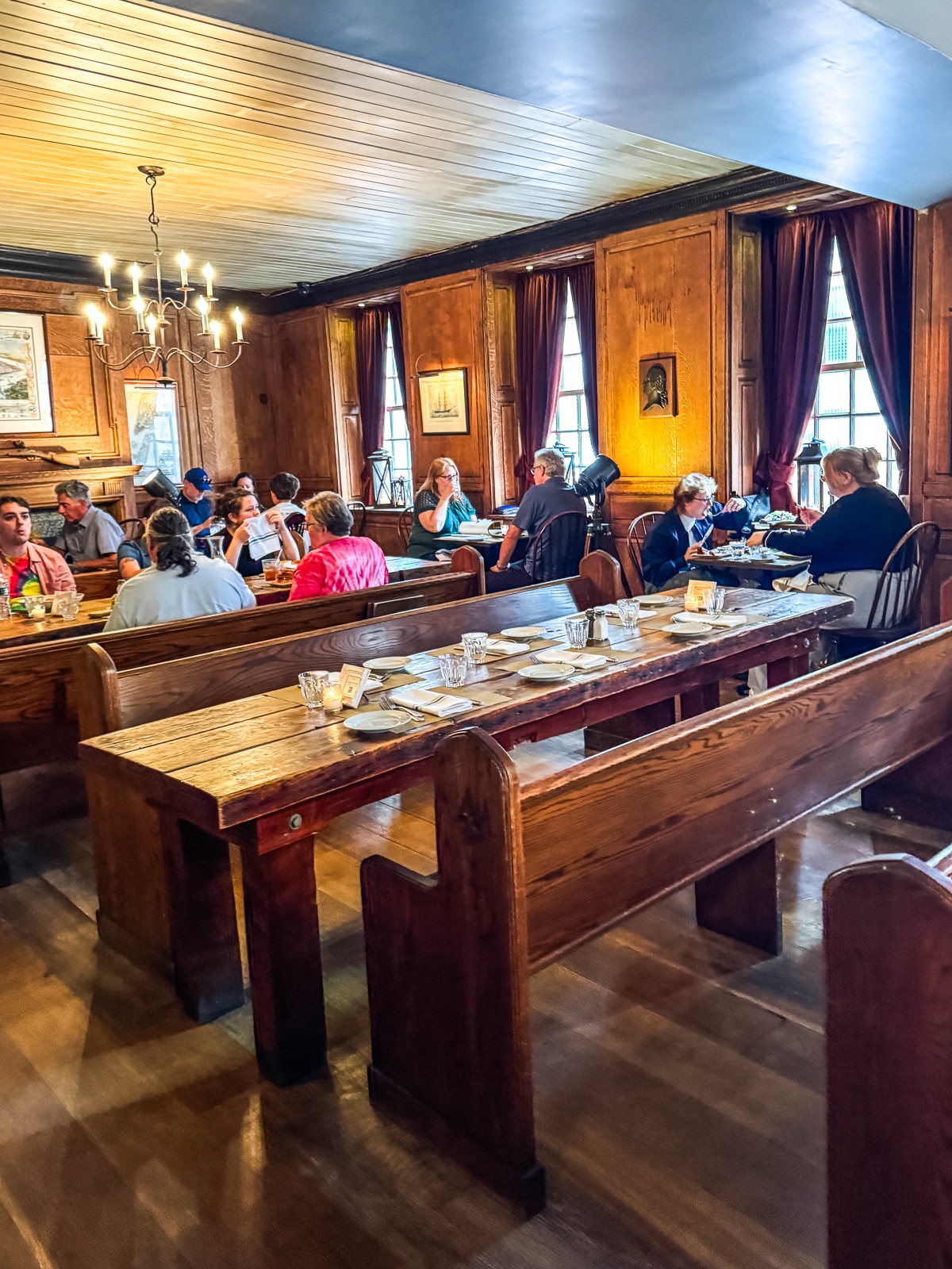 Groups of people sit at wooden tables and benches in a warmly lit dining room with wood-paneled walls, large windows, and a chandelier.