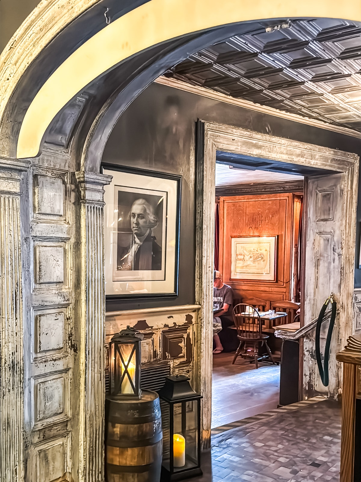 A rustic interior with aged wooden archways, a lantern on a barrel, and framed portraits on the walls; a person sits at a table in the adjoining room.