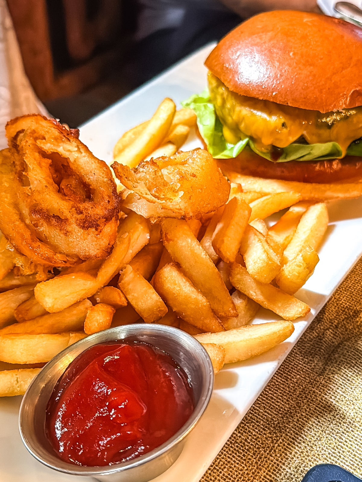 A cheeseburger with lettuce on a bun, served with French fries, onion rings, and a metal cup of ketchup on a rectangular white plate.