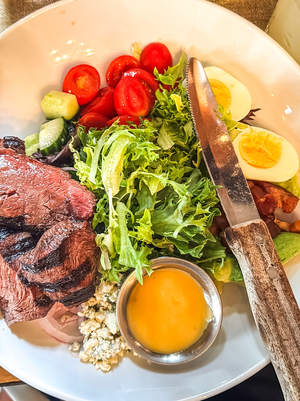 A plate with grilled steak slices, mixed greens, cherry tomatoes, cucumber, hard-boiled egg, bleu cheese crumbles, and a small cup of dressing. A knife rests on the plate.