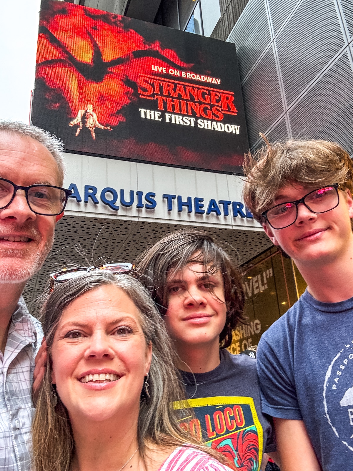 Four people stand in front of the Marquis Theatre with a digital sign for "Stranger Things: The First Shadow" displayed above them.