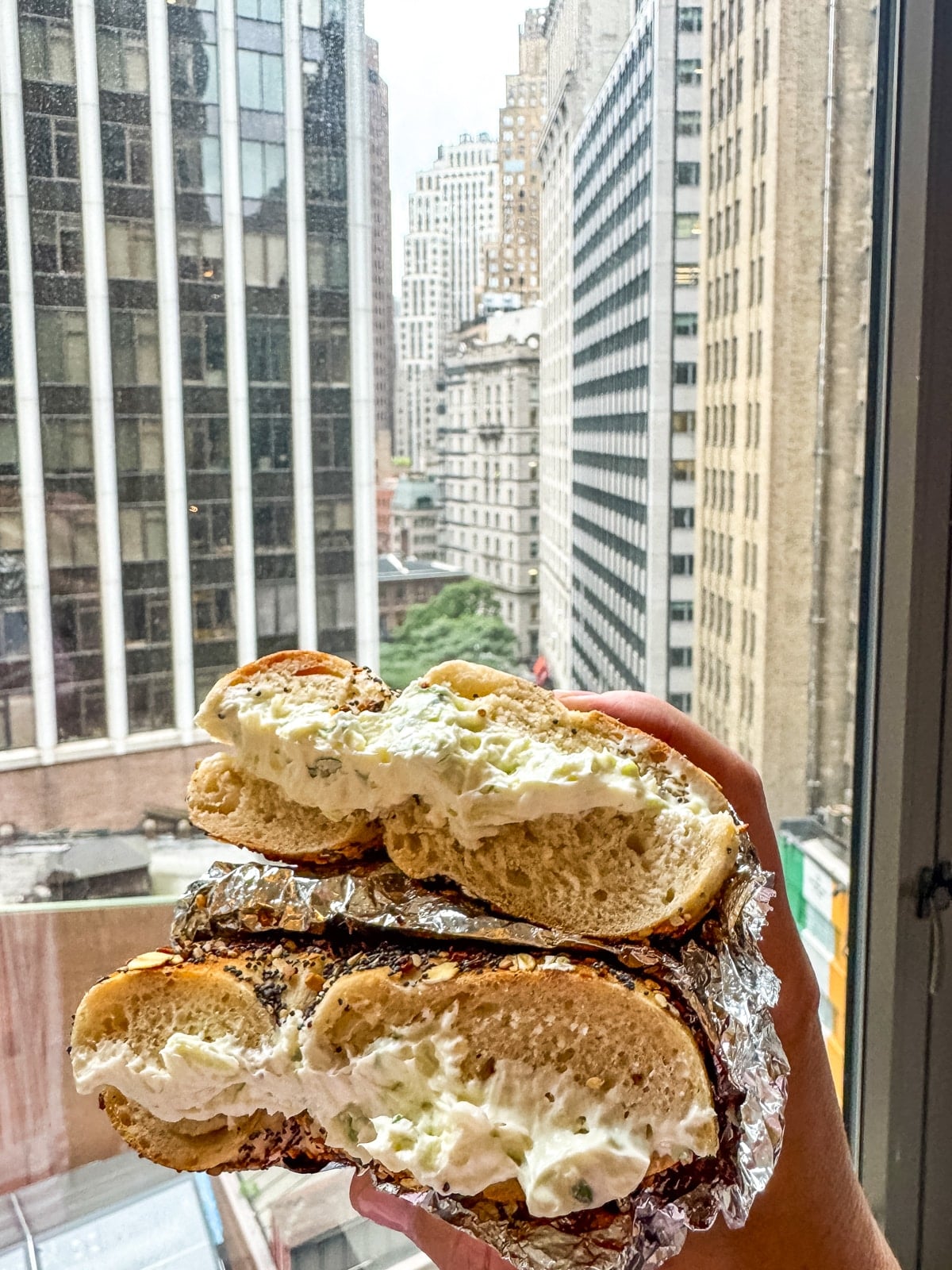 A hand holds a halved, cream cheese-filled bagel wrapped in foil in front of a window with a view of tall city buildings.