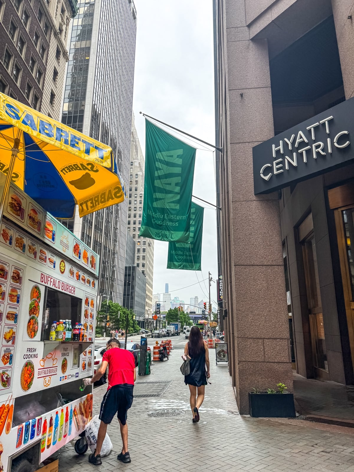 A man stands at a Sabrett hot dog cart on a city sidewalk near the Hyatt Centric hotel; a woman walks away down the street.