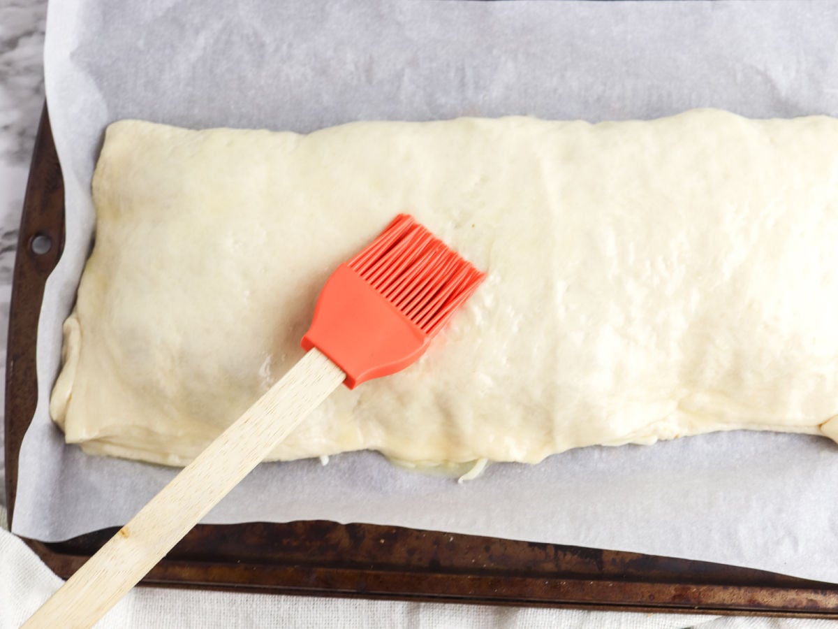 A pastry brush with a red silicone head is being used to apply an egg wash to an unbaked Chicken Parm Stromboli on a parchment-lined baking sheet.