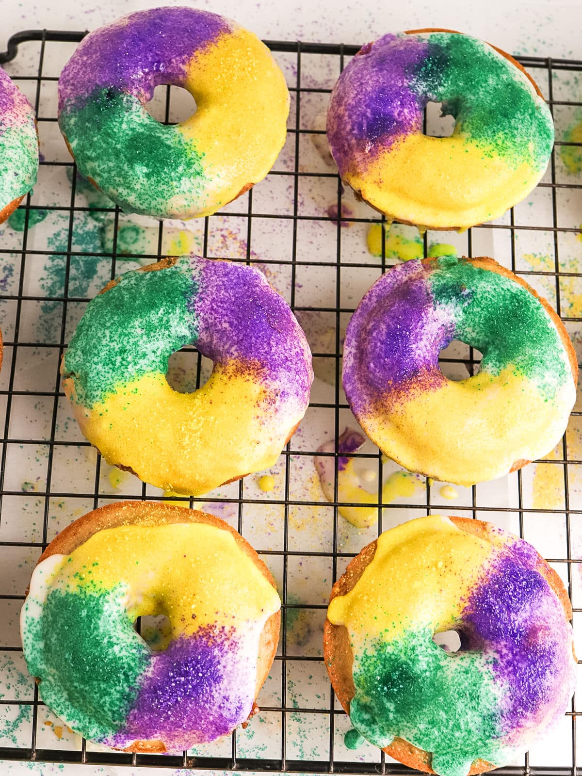 Six donuts with white icing are decorated with green, yellow, and purple sugar, arranged on a black wire cooling rack. Some colored sugar is scattered on the surface.