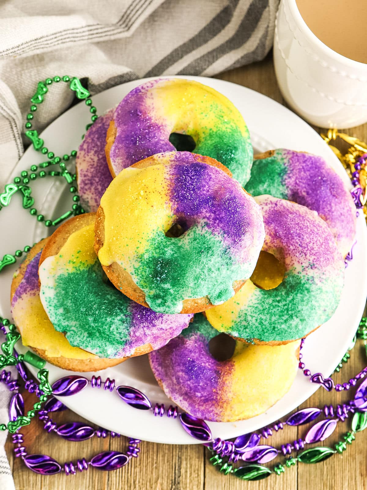 A plate of donuts decorated with yellow, green, and purple icing, surrounded by Mardi Gras beads, next to a cup on a wooden surface.