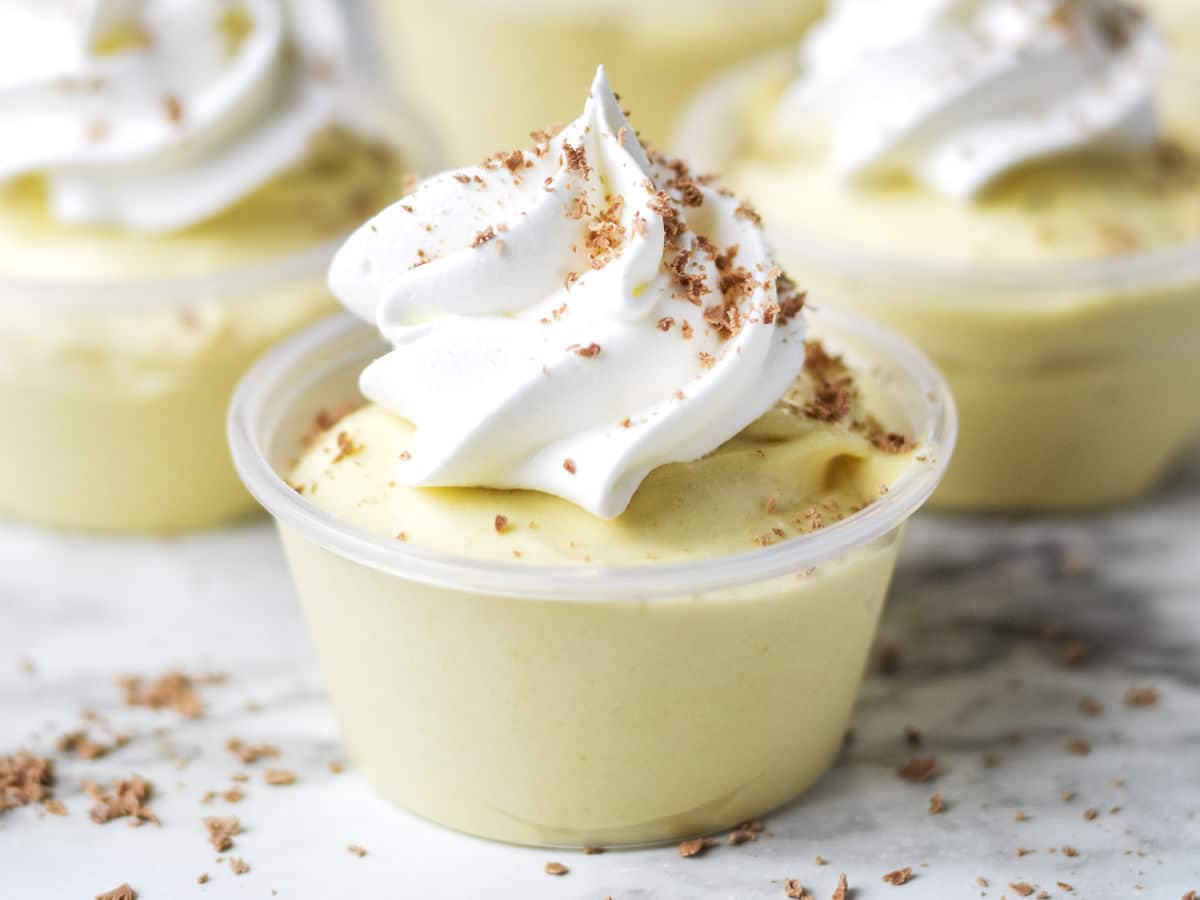 A small plastic cup filled with yellow mousse, topped with whipped cream and chocolate shavings, sits on a marble surface.
