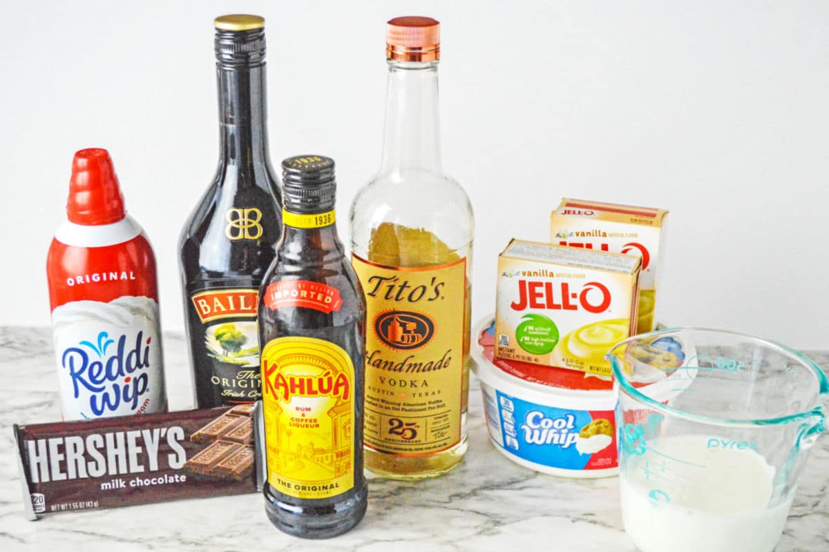 Various ingredients on a countertop, including Reddi Wip, Baileys, Kahl&uacute;a, Tito's vodka, Jell-O vanilla pudding mixes, Cool Whip, Hershey's chocolate, and a measuring cup with milk.