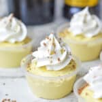 Close-up of small plastic cups filled with yellow pudding, topped with whipped cream and chocolate shavings, arranged on a light surface.