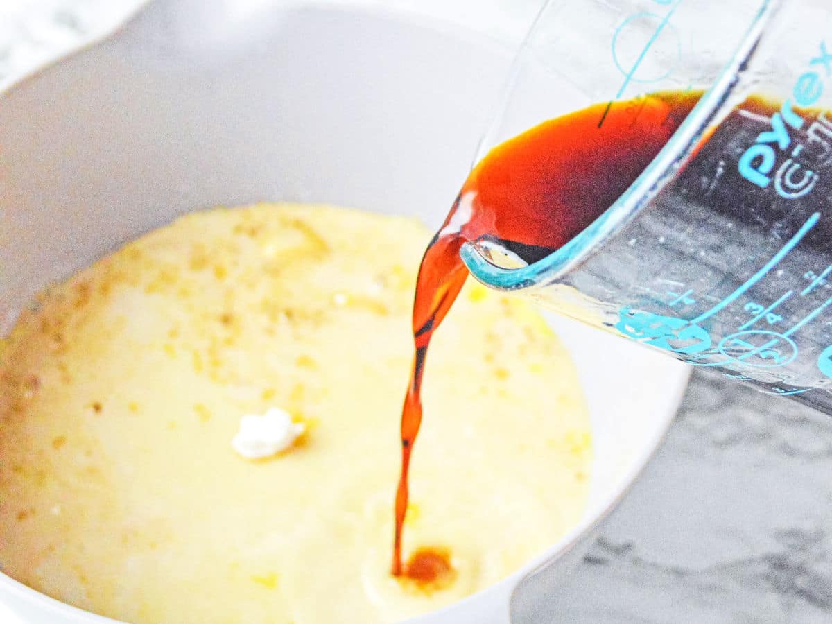 A measuring cup pours a dark liquid, likely vanilla extract, into a bowl of light-colored batter on a countertop.