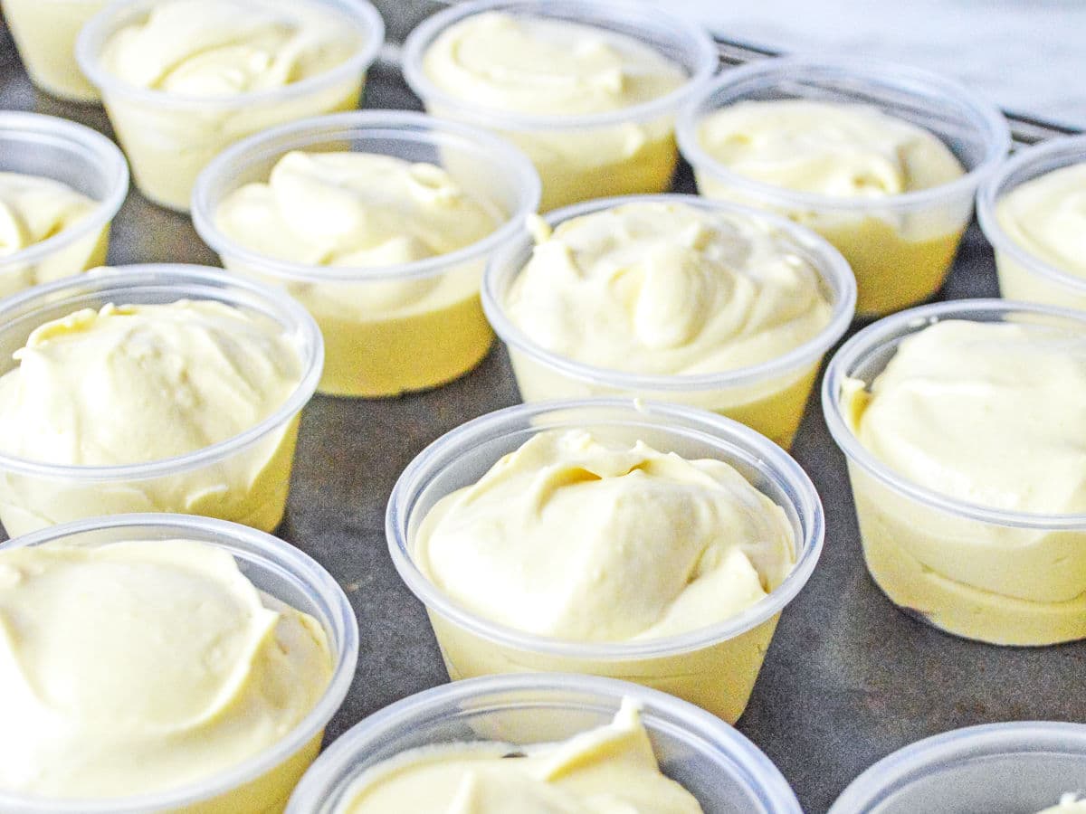 Several small clear plastic cups filled with creamy yellow pudding or mousse are arranged closely together on a flat surface.