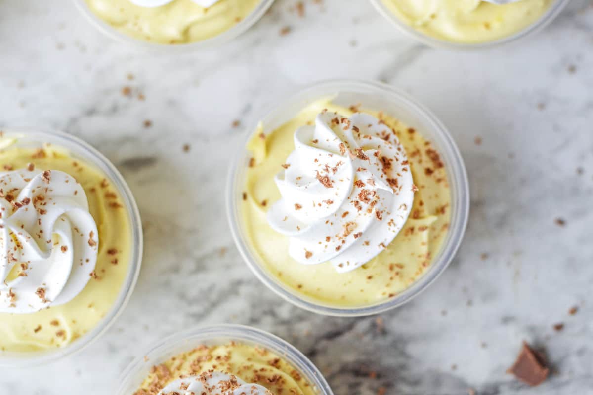 Small cups filled with yellow mousse, each topped with a swirl of whipped cream and sprinkled with chocolate shavings, set on a light marble surface.