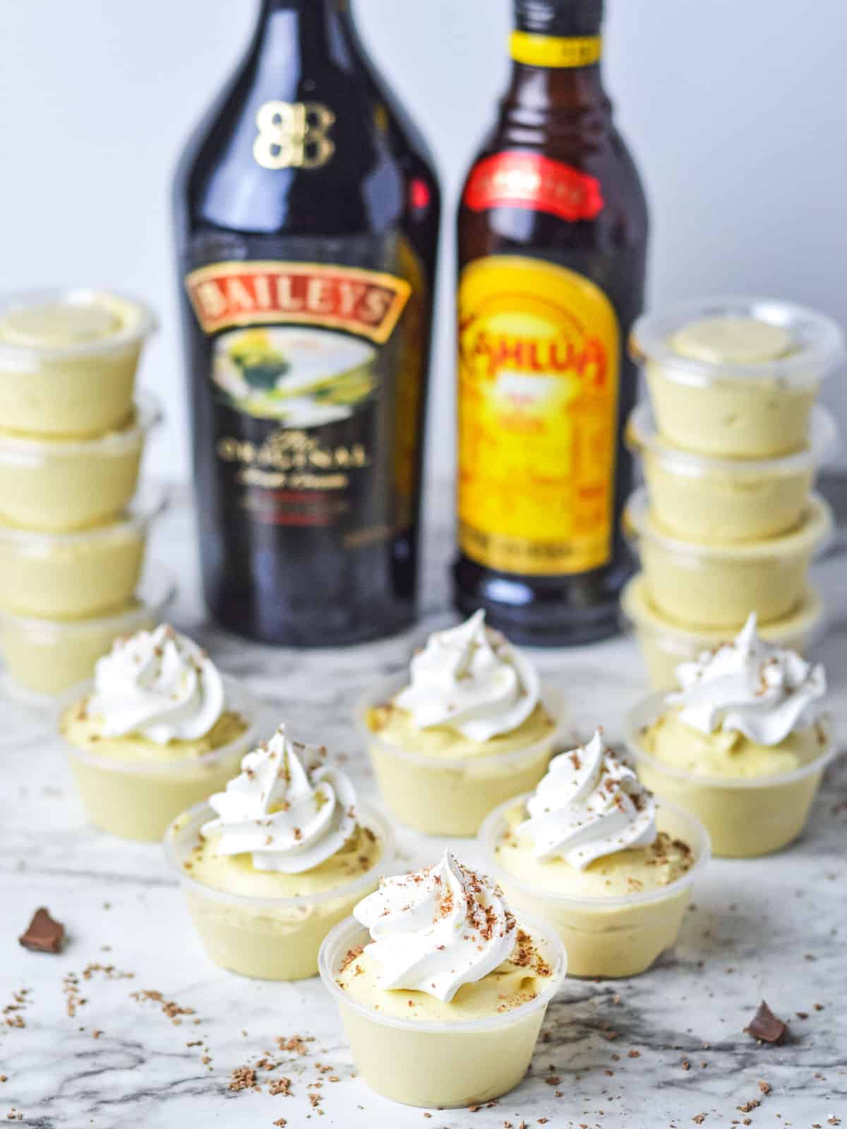 Mini dessert cups topped with whipped cream are arranged in front of Baileys and Kahlua bottles, with more dessert cups stacked in the background on a marble surface.