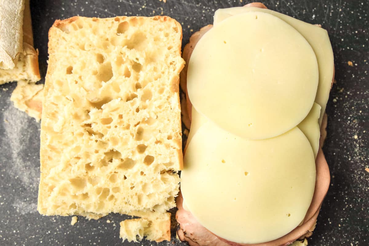 Open sandwich with two slices of provolone cheese and deli meat on a slice of bread, next to the top piece of bread, on a dark surface.