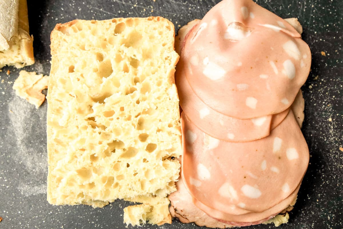 A close-up of an open sandwich with airy ciabatta bread on the left and several slices of mortadella sausage on the right, placed on a dark surface.