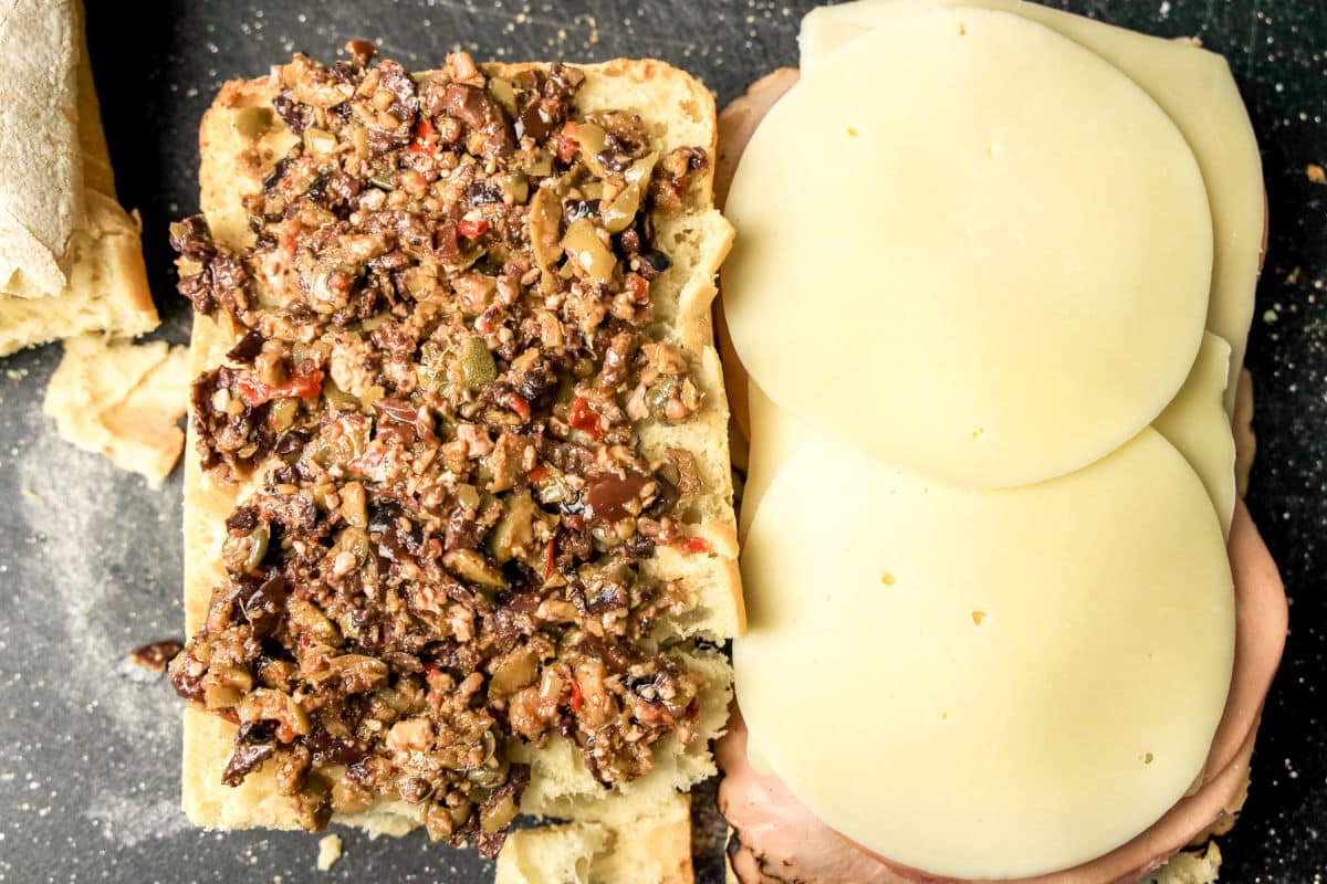 Open sandwich with olive tapenade on one half of ciabatta bread and stacked slices of provolone cheese and deli meat on the other half, on a dark surface.