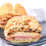 A sliced ciabatta sandwich filled with layers of deli meat, cheese, and chopped olive salad, served on a dark plate.