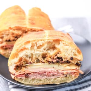 A sliced ciabatta sandwich filled with layers of deli meat, cheese, and chopped olive salad, served on a dark plate.