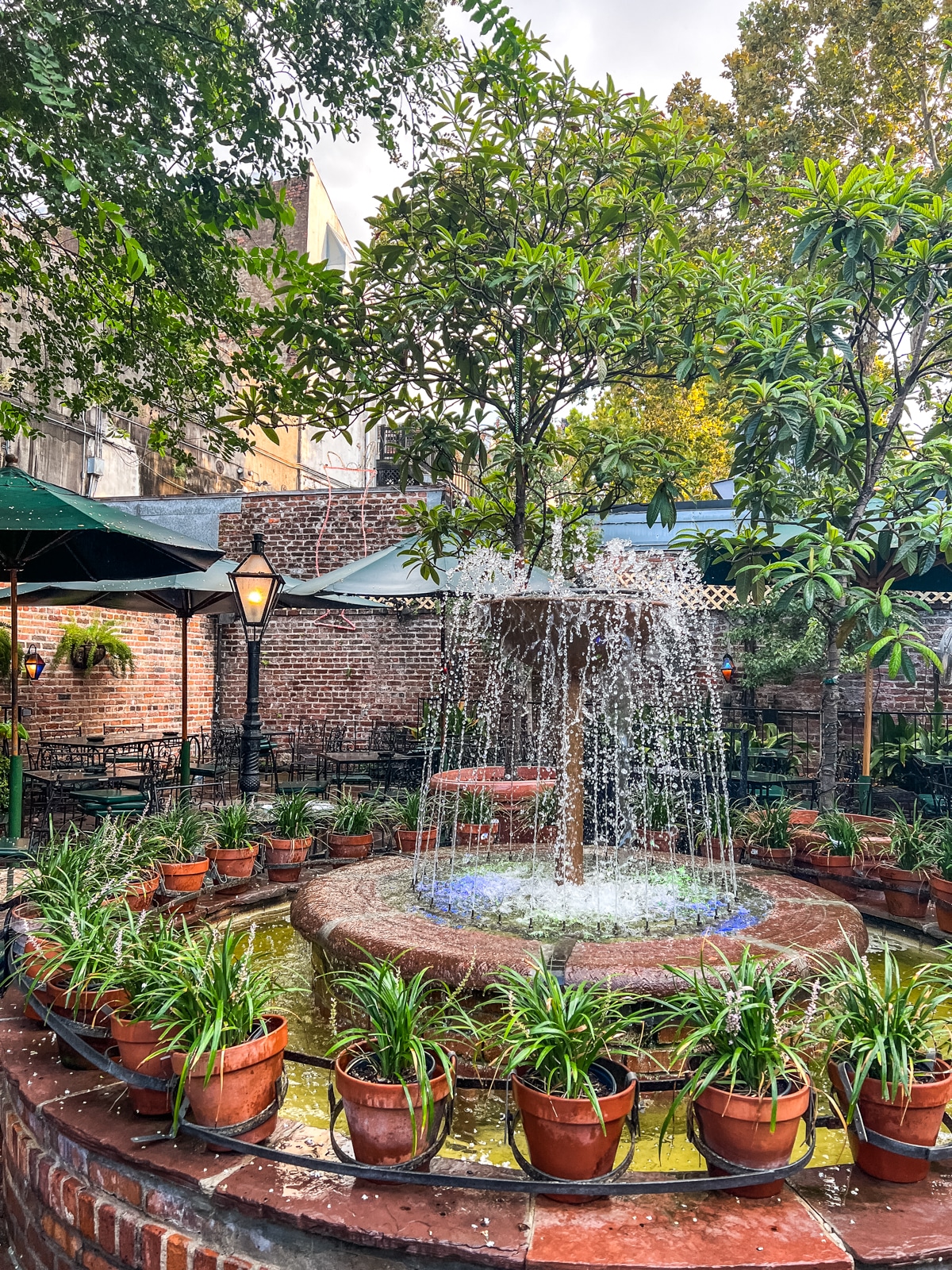 A brick courtyard fountain surrounded by potted green plants, with outdoor tables, chairs, and a brick wall in the background.