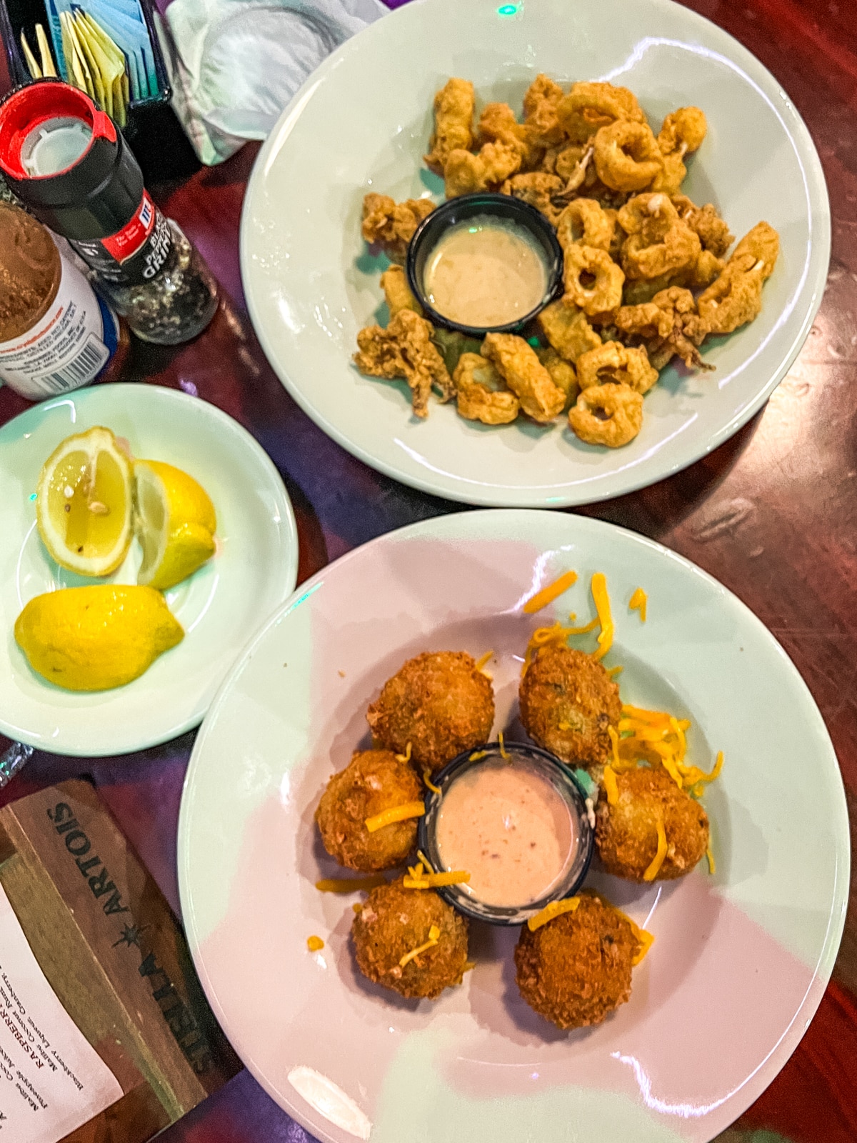 Two plates of fried appetizers: one with fried calamari and dipping sauce, another with fried cheese balls and dipping sauce. A small plate with lemon wedges is beside them.
