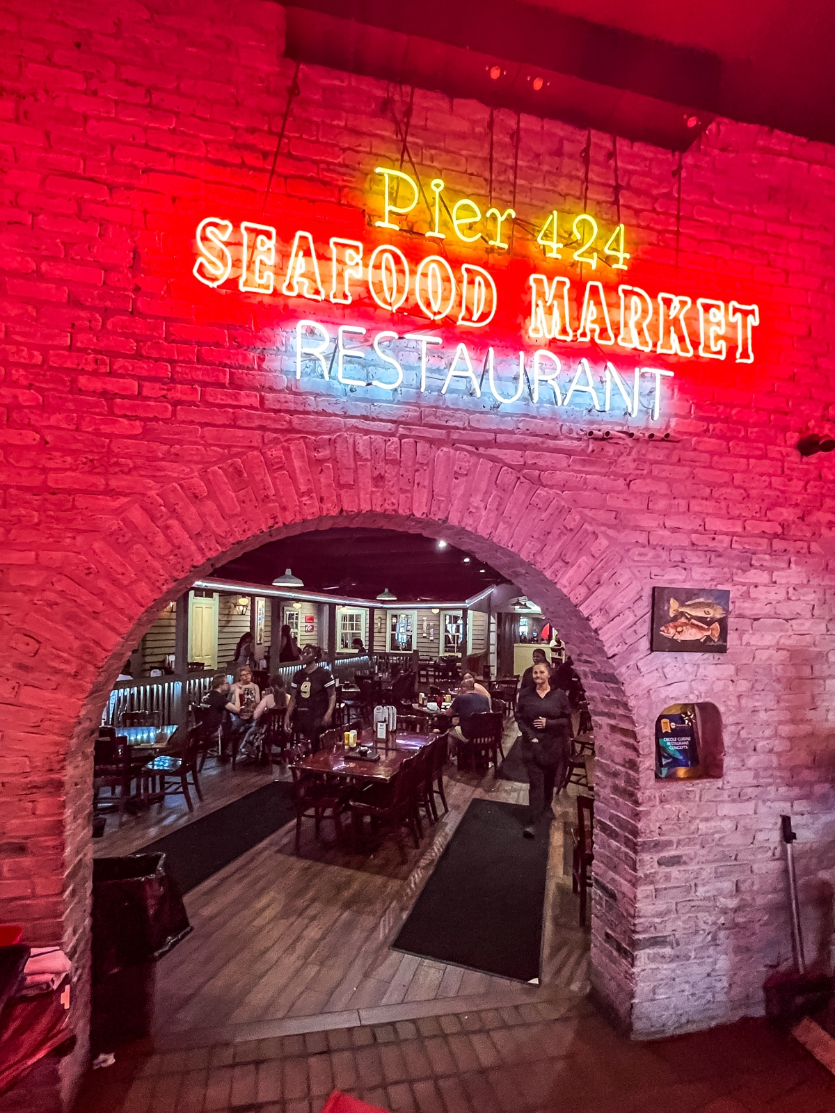 Neon sign for "Pier 424 Seafood Market Restaurant" above a brick archway entrance, with diners and tables visible inside the restaurant.