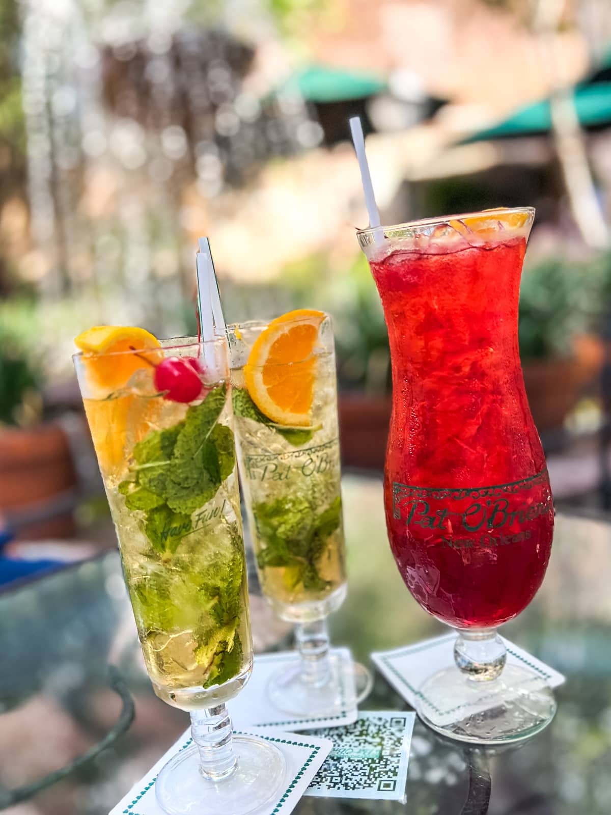 Three tall glasses with cocktails, two with mint, orange, and cherry garnishes, and one with a bright red drink and orange slice, placed on a table with outdoor background.
