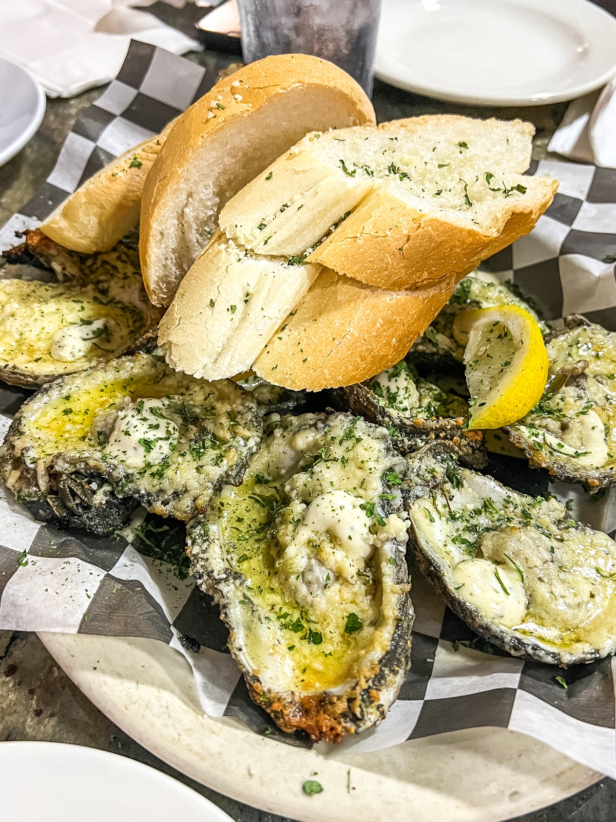 A plate of baked oysters topped with herbs and butter, served with slices of toasted bread and a lemon wedge on checkered paper.