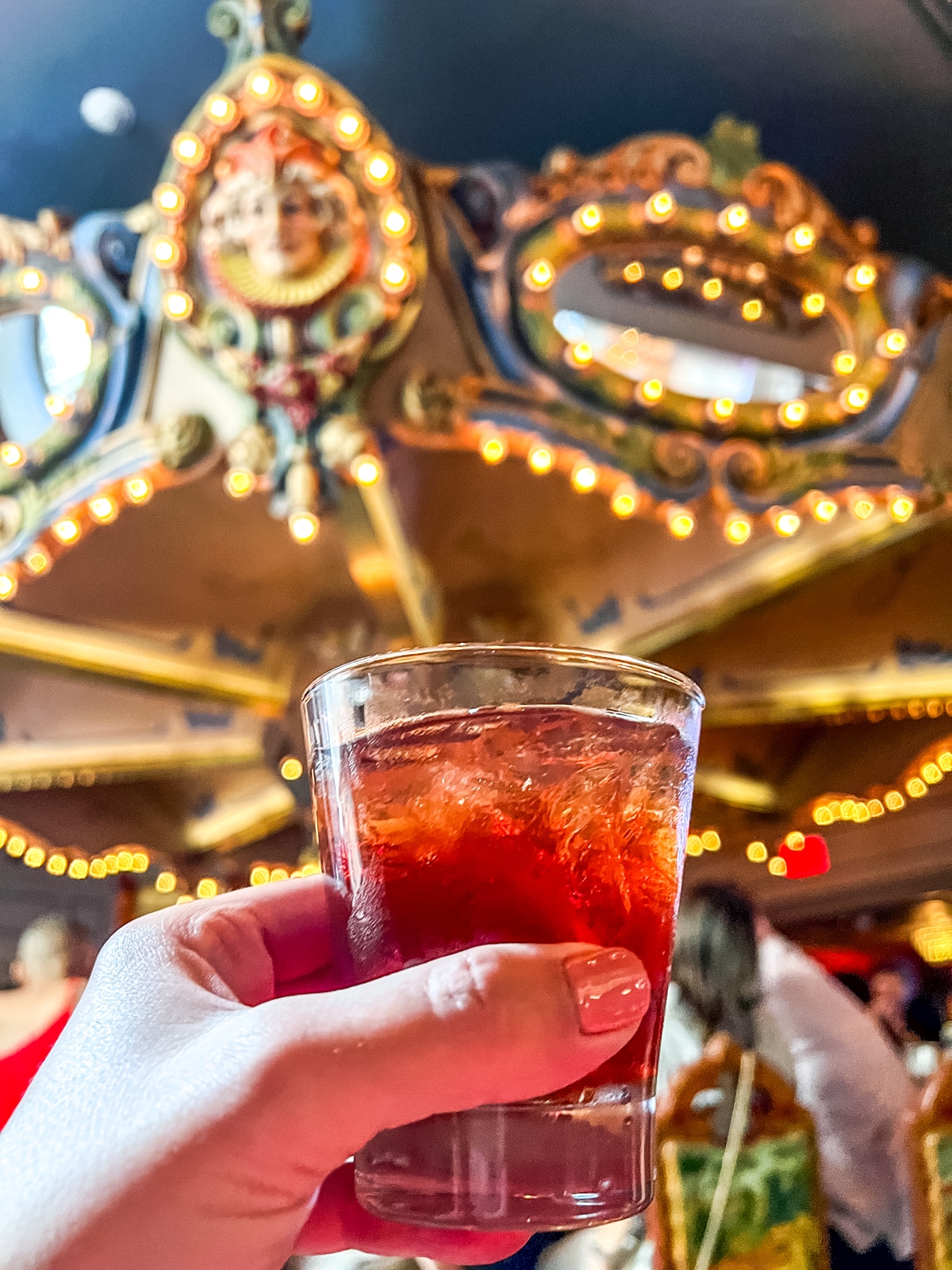 A hand holds a glass of a red cocktail in front of a carousel-themed ceiling with glowing lights and ornate decorations.