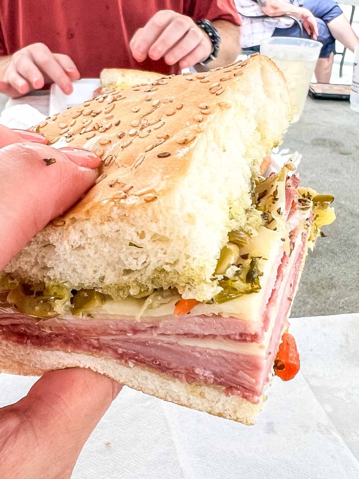 A hand holding a thick sandwich with sesame seed bread, layers of meat, cheese, and chopped vegetables; people sitting at a table in the background.