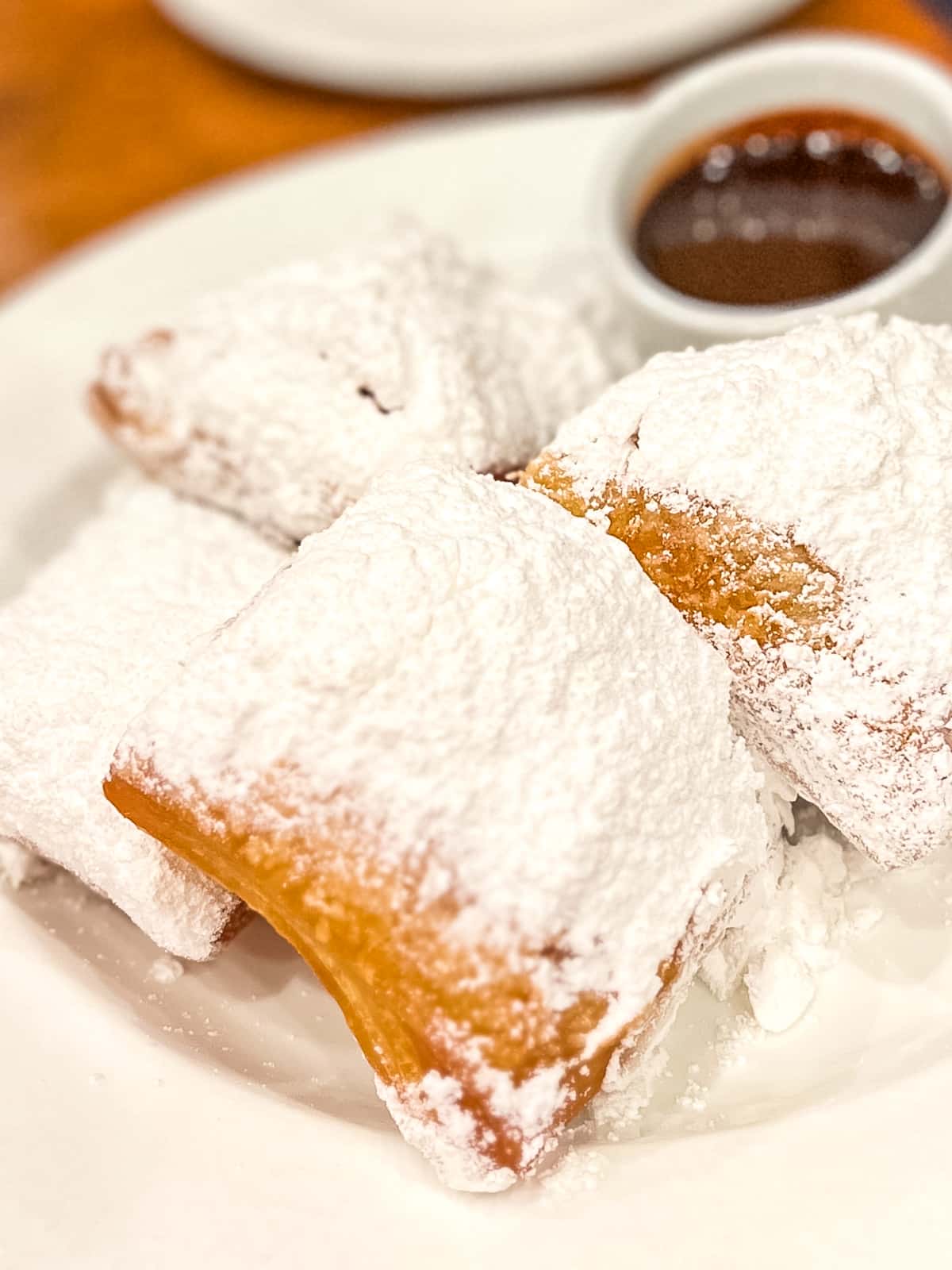 Three powdered sugar-covered beignets are served on a white plate with a small cup of chocolate dipping sauce in the background.