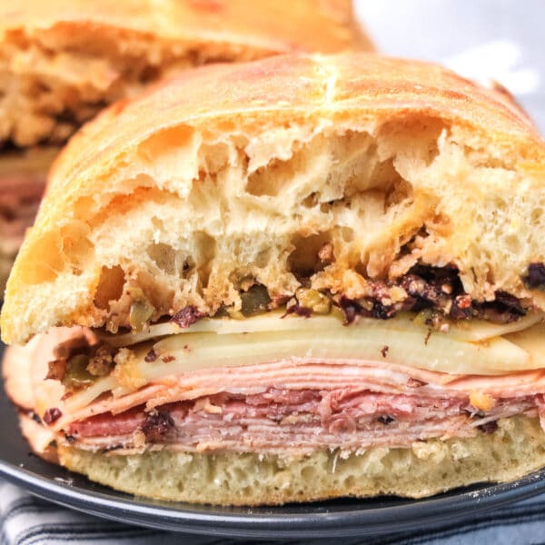 A sandwich with layers of deli meat, cheese, and chopped olives on crusty bread, partially eaten, sits on a black plate.