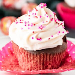 A chocolate cupcake in a pink polka dot wrapper topped with white frosting and pink, red, and purple sprinkles.