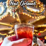 A hand holding a cocktail in front of a brightly lit, carousel-themed bar, with the text "What to Eat in New Orleans" at the top.