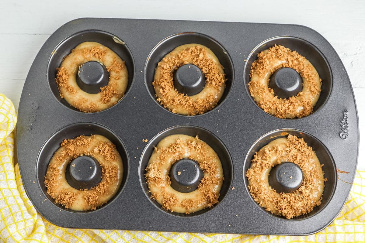 A six-cavity donut pan filled with unbaked donut batter, each topped with a crumbly streusel, sits on a white surface next to a yellow kitchen towel.
