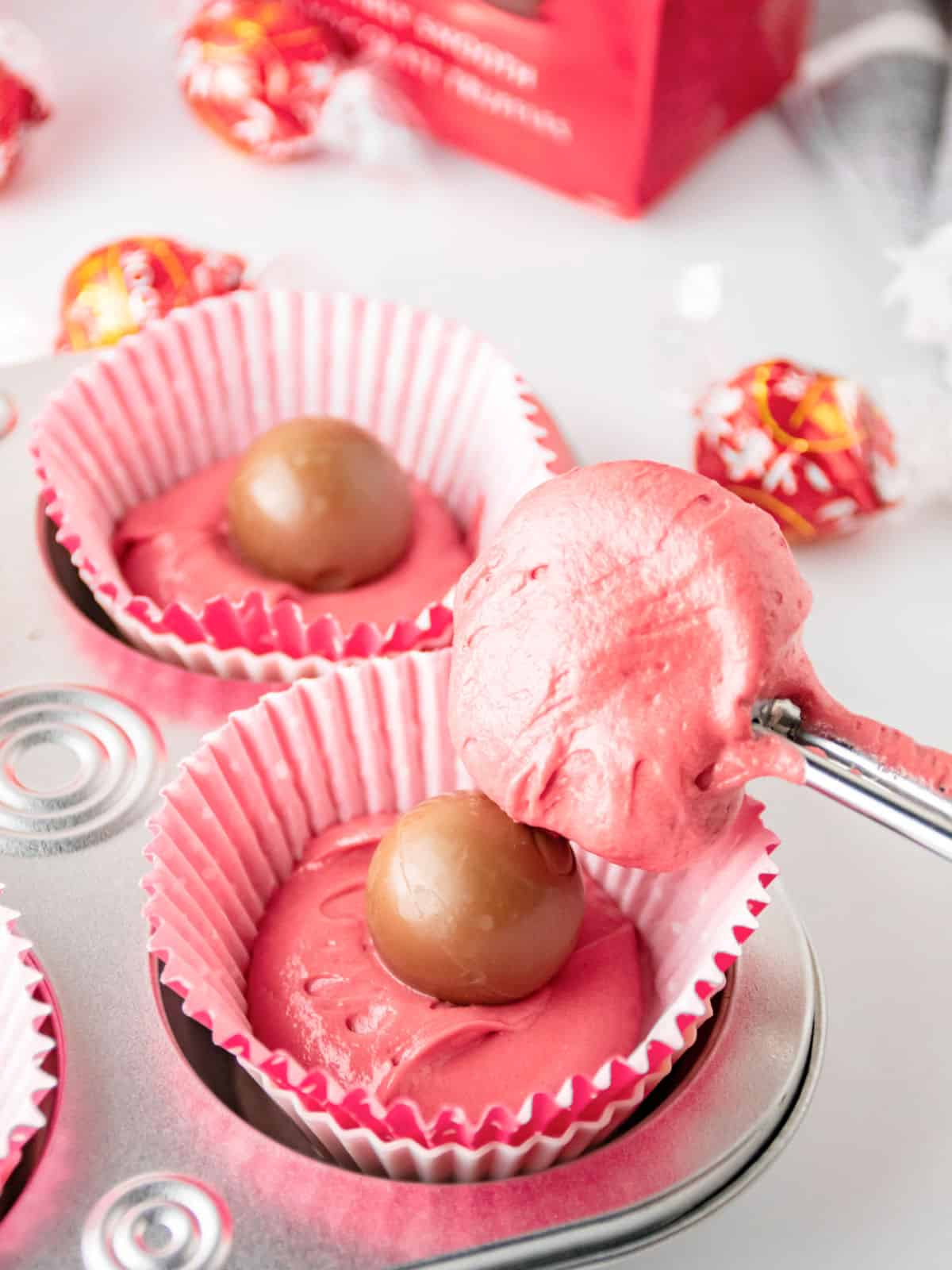 A muffin tin lined with pink paper cups, filled with pink batter and a chocolate truffle in the center, with a scoop of batter being added to one cup.