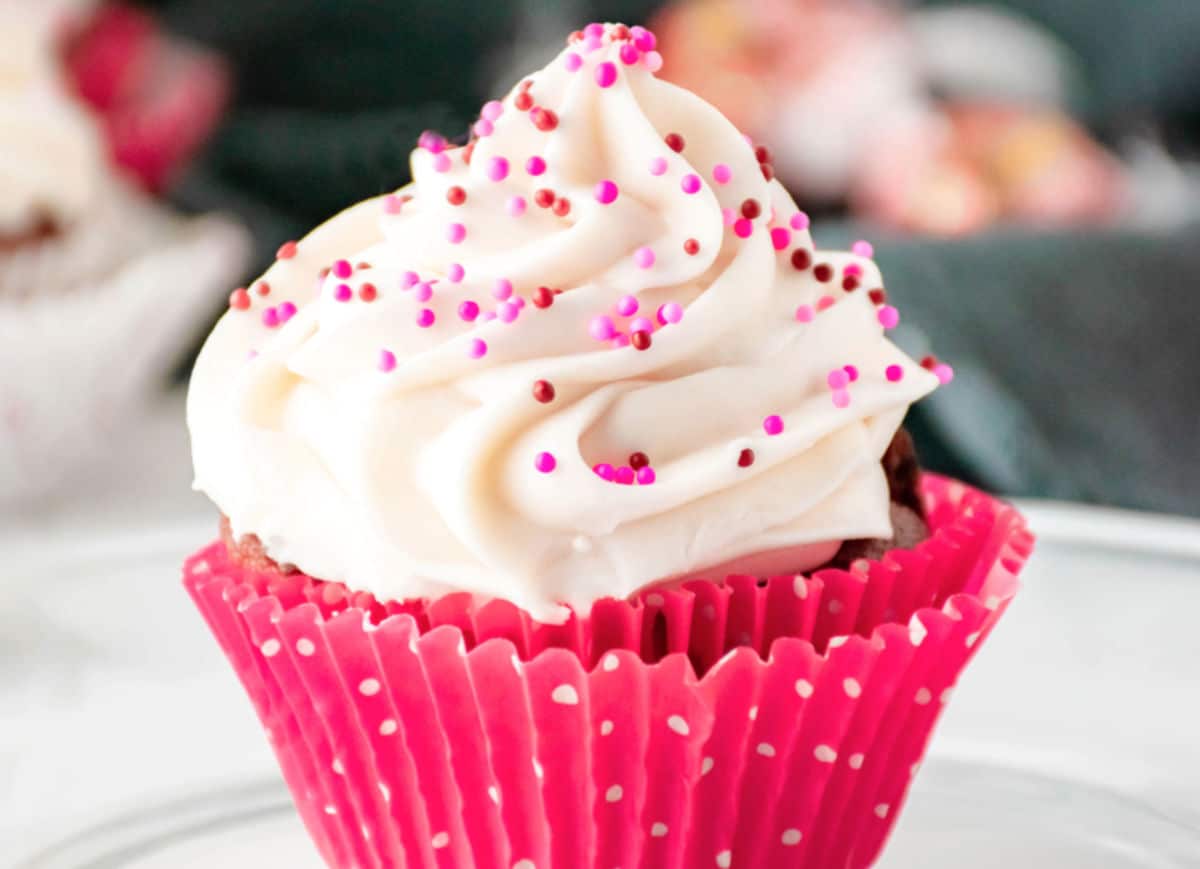A cupcake in a pink polka dot wrapper topped with white frosting and pink and red sprinkles.