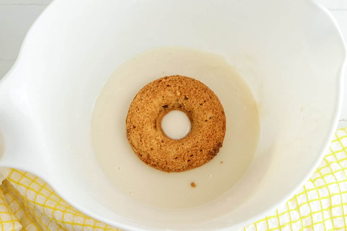 A round, baked donut sits in a shallow layer of liquid inside a white mixing bowl, with a yellow checkered cloth partially visible underneath.