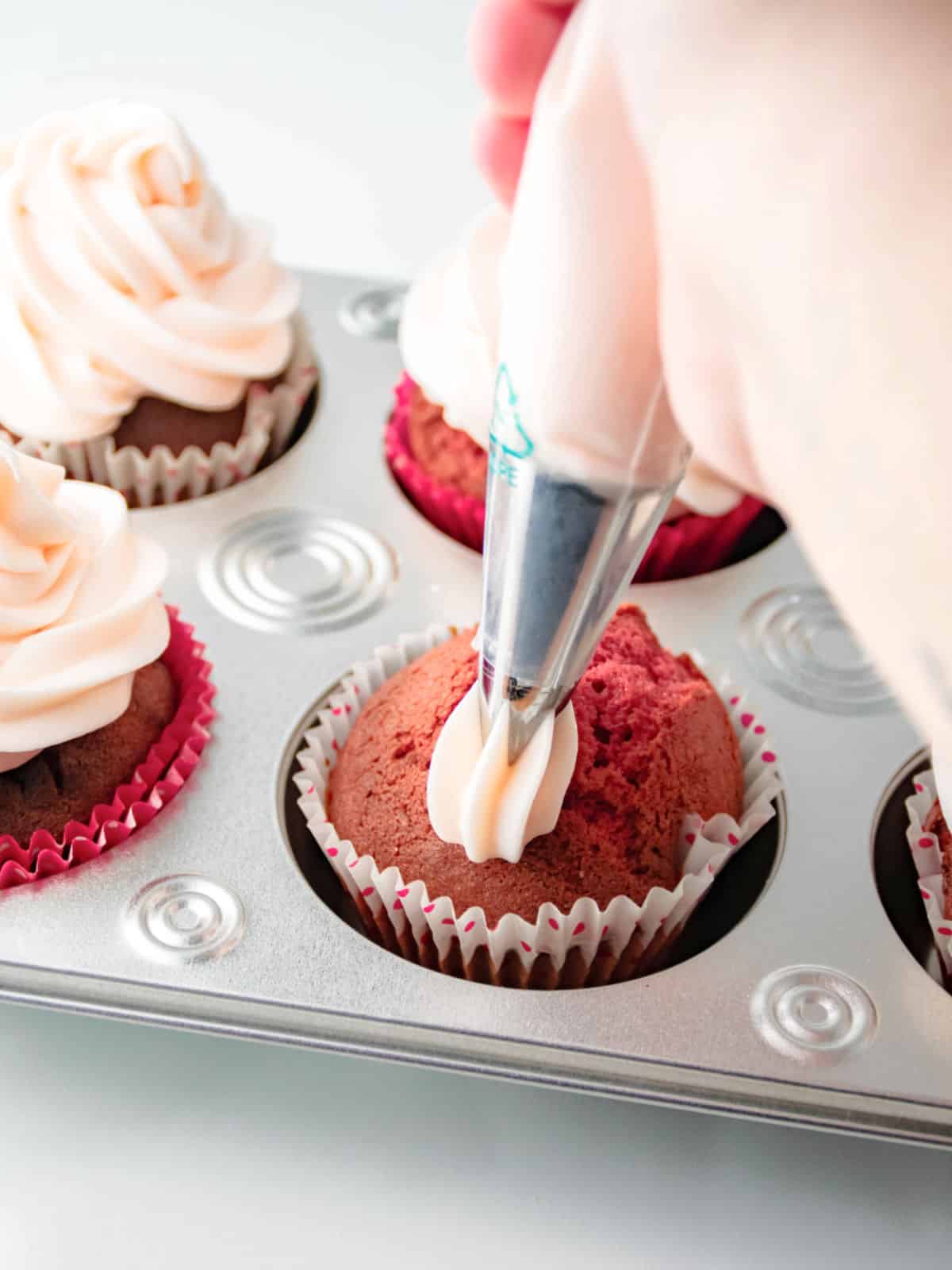 A hand is piping frosting onto a cupcake in a muffin tray, with several already frosted cupcakes nearby.