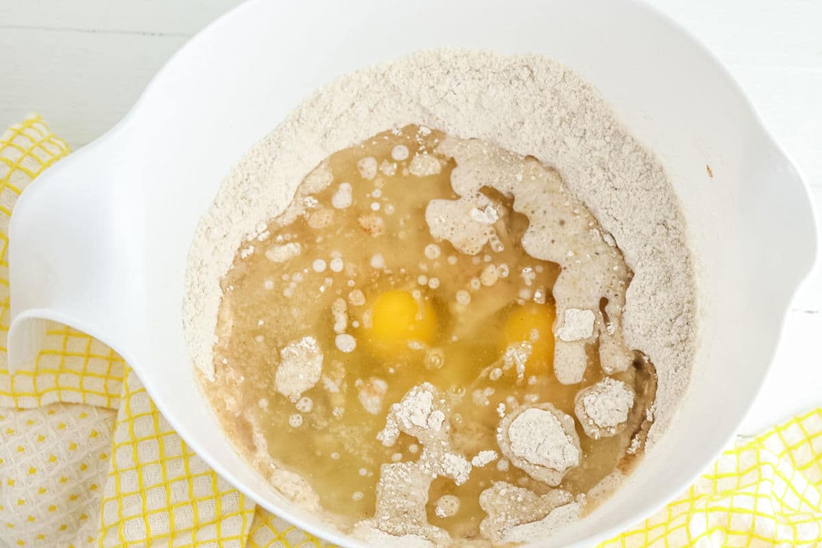 A mixing bowl containing flour, eggs, and liquid ingredients before being mixed, placed on a yellow and white towel.