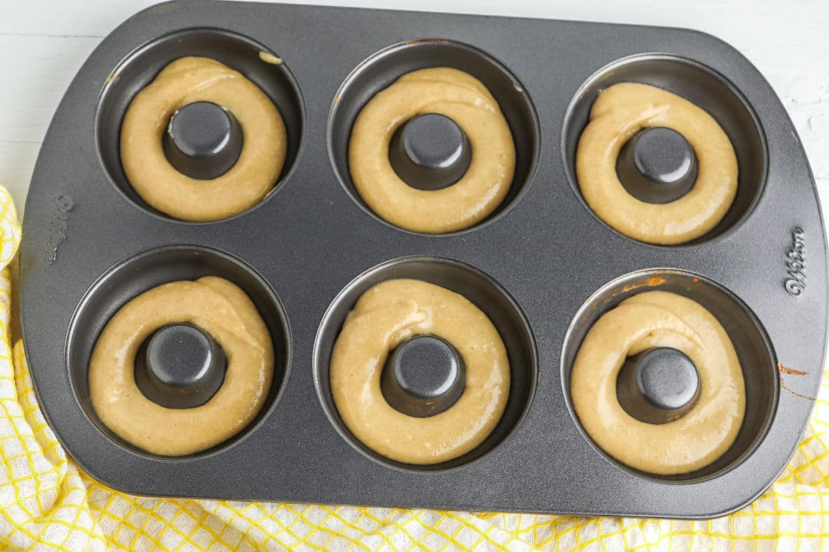 A donut pan filled with raw donut batter sits on a white surface beside a yellow checkered cloth.