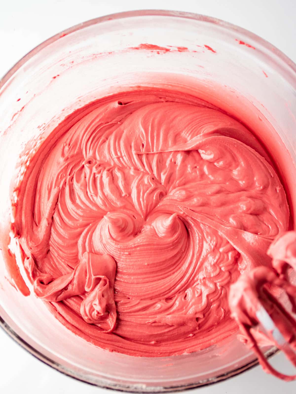 A glass mixing bowl filled with smooth, whipped pink frosting, with a bit of frosting on a metal beater in the foreground.