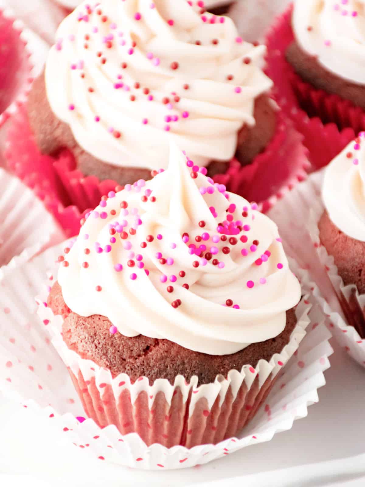 Red velvet cupcakes in pink paper liners topped with swirls of white frosting and pink and red sprinkles.