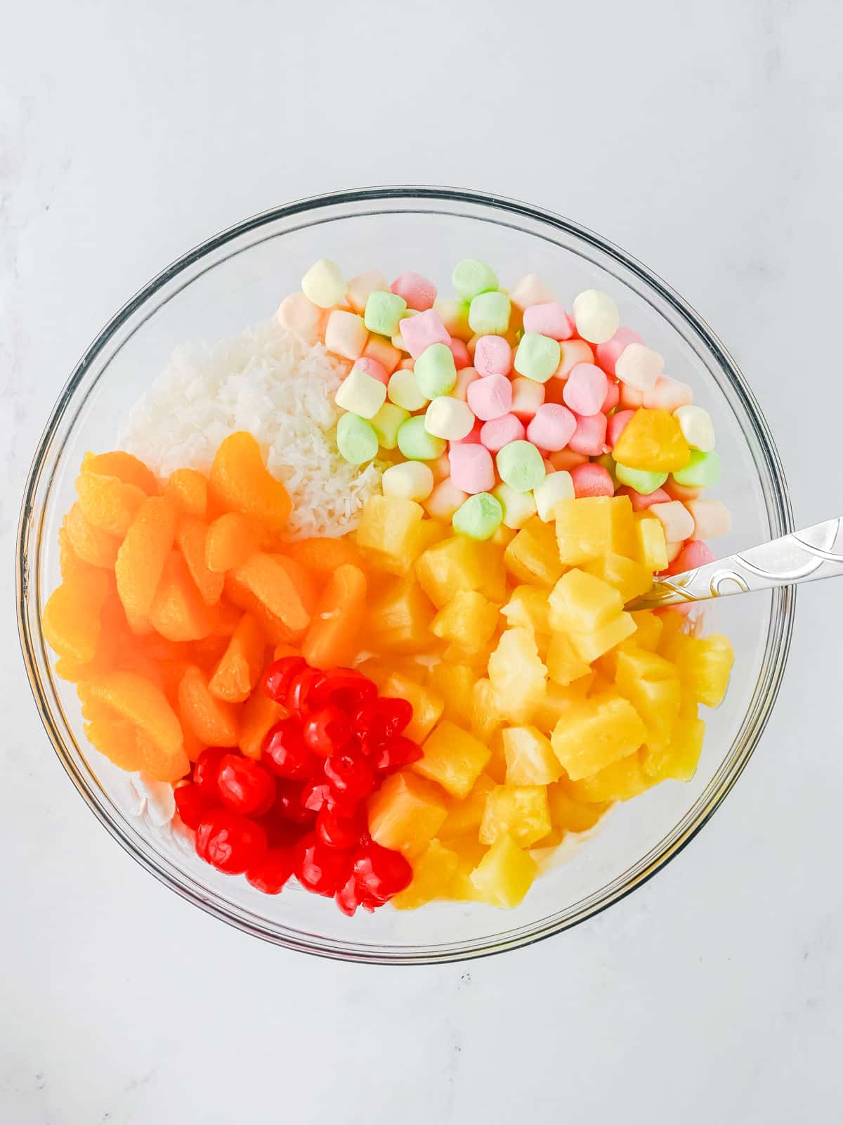 A glass bowl containing colorful mini marshmallows, shredded coconut, pineapple chunks, mandarin orange segments, and maraschino cherries. A spoon rests on the side of the bowl.