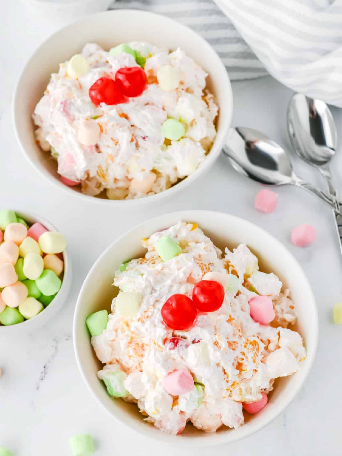 Two bowls of fruit salad topped with whipped cream, marshmallows, and cherries, with spoons and extra marshmallows on the side.