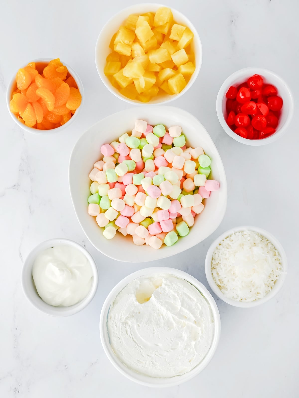 Bowls containing mini marshmallows, whipped topping, shredded coconut, maraschino cherries, pineapple chunks, and mandarin oranges on a white surface.